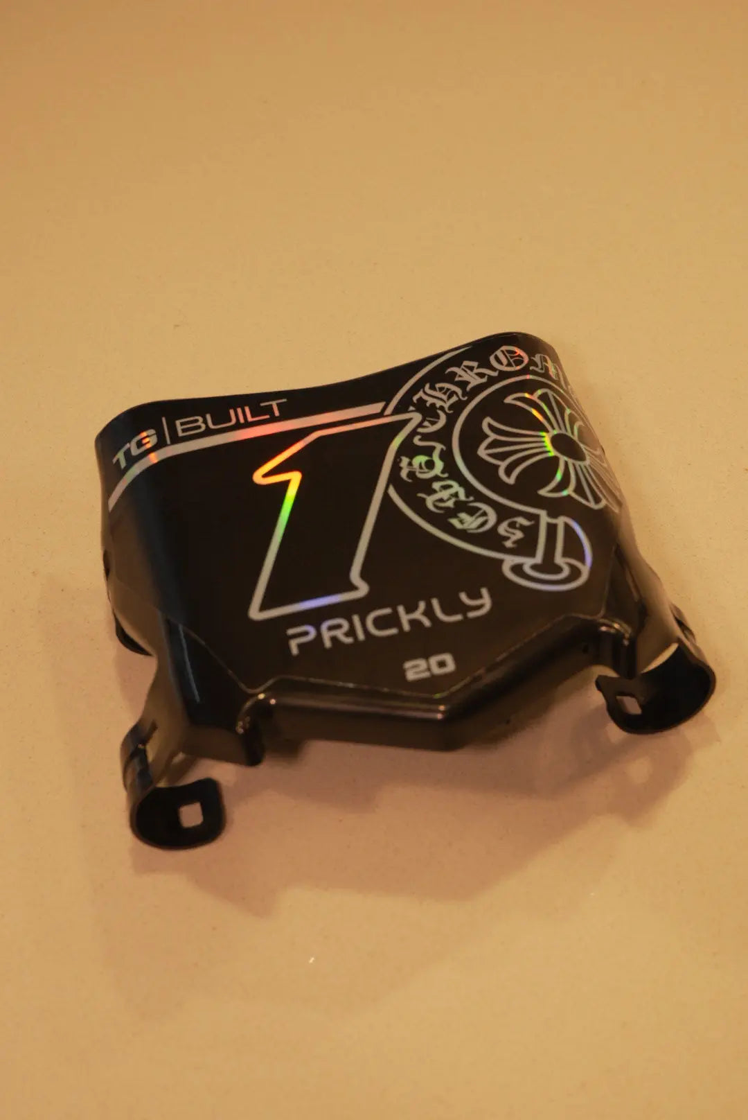 A black custom motorcycle helmet shell or fairing piece with graphic decals, including a large holographic '1' and 'PRICKLY' text, photographed on a plain beige surface.