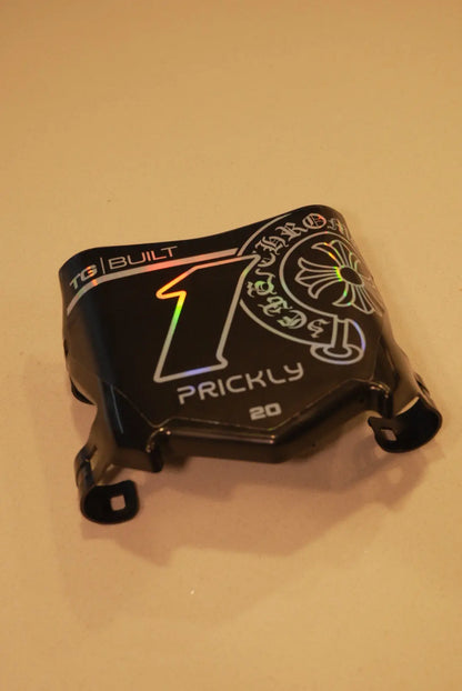 A black custom motorcycle helmet shell or fairing piece with graphic decals, including a large holographic '1' and 'PRICKLY' text, photographed on a plain beige surface.